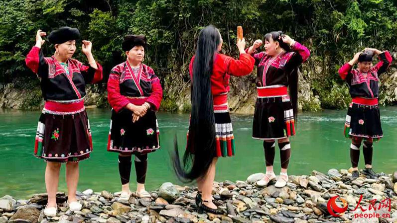 行進(jìn)中國(guó)丨“長(zhǎng)發(fā)”引客來(lái)雨后初晴，在廣西龍勝各族自治縣黃洛瑤寨的小河邊，幾名紅瑤婦女有說(shuō)有笑地梳洗著如瀑布般傾瀉的長(zhǎng)發(fā)。伴隨著流水聲，她們時(shí)不時(shí)齊聲合唱著《長(zhǎng)發(fā)謠》，將長(zhǎng)發(fā)盤(pán)于頭頂，再配上精美的發(fā)飾，形成“烏龍盤(pán)發(fā)”。